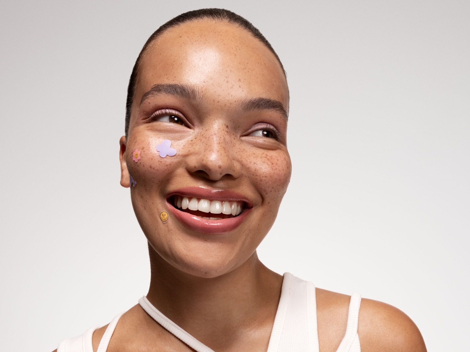 Eine lächelnde Frau mit Sommersprossen trägt bunte Gesichtsaufkleber in Form von Blumen und einem Smiley, mit glänzenden Lippen und leichtem Lidschatten.