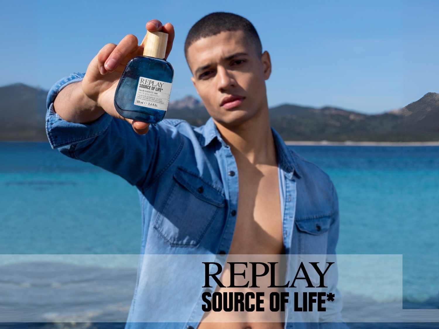 Een man houdt een blauw flesje Replay Source of Life eau de toilette vast tegen een achtergrond van blauwe zee en lucht.