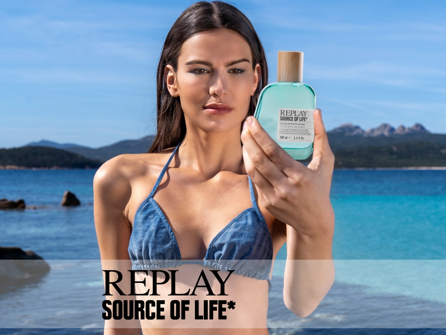Een vrouw in bikini houdt een flesje Replay Source of Life parfum vast tegen een achtergrond van zee en blauwe lucht, met de tekst "REPLAY SOURCE OF LIFE*".