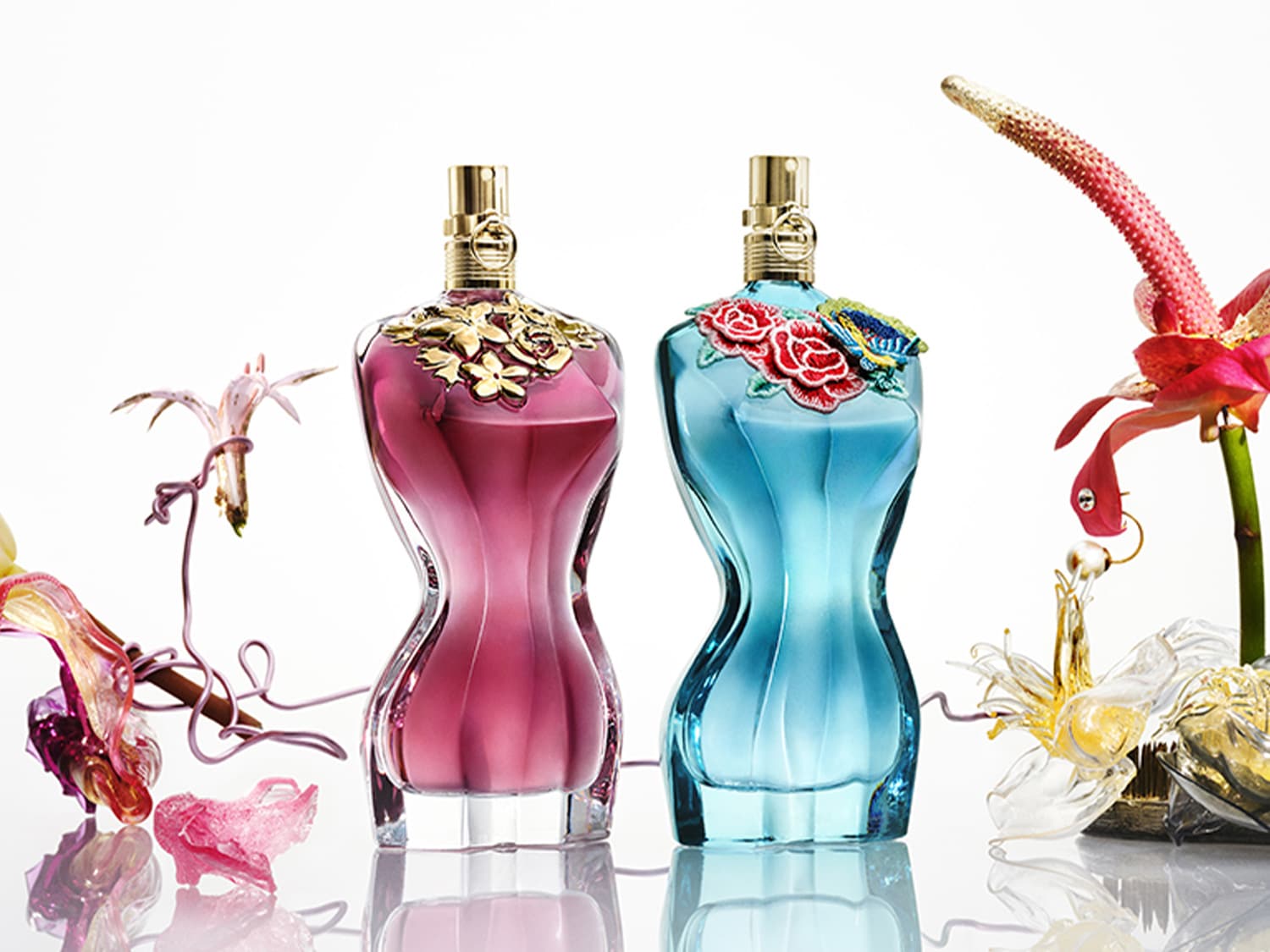 Twee Jean Paul Gaultier parfumflessen, de ene roze met gouden bloemendetails en de andere blauw met rode rozen en een blauwe vlinder, omringd door bloemen.