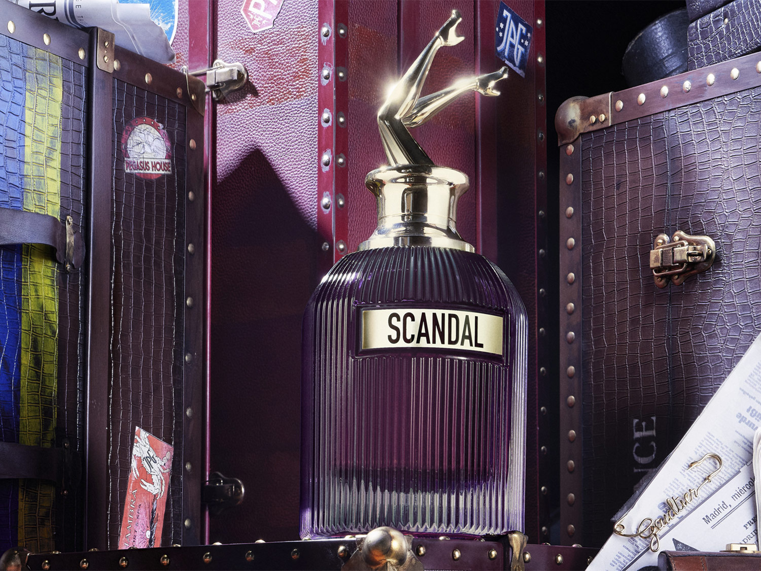 De Scandal parfumfles, gehuld in een dieppaarse tint, staat trots tussen vintage koffers, bekroond met een gouden sculptuur van benen in de lucht.