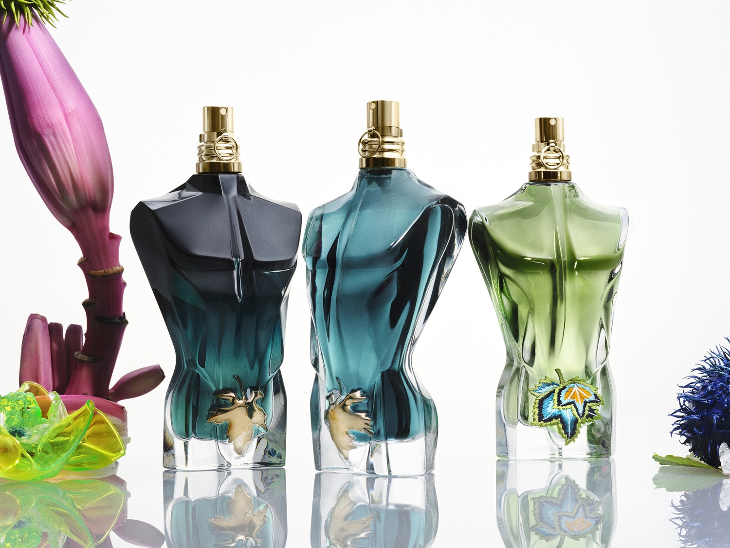 Drie flessen Jean Paul Gaultier Le Beau parfum, elk in de vorm van een mannelijk torso, in zwart, blauw en groen, met gouden accenten en bloemdecoraties.