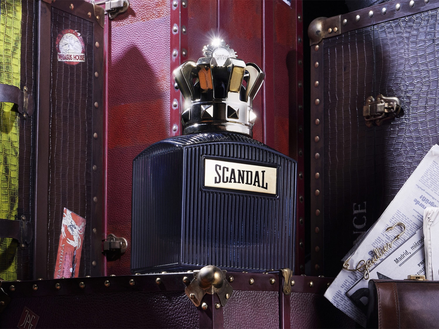 Een zwarte Scandal parfumfles met een gouden kroon als dop, geplaatst tussen vintage koffers en kranten.