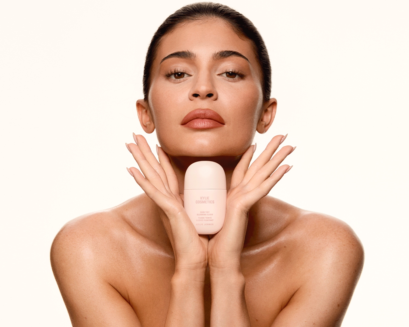 Une femme tient un flacon rose de Kylie Cosmetics Skin Tint Blurring Elixir, promettant un éclat doux et uniforme.