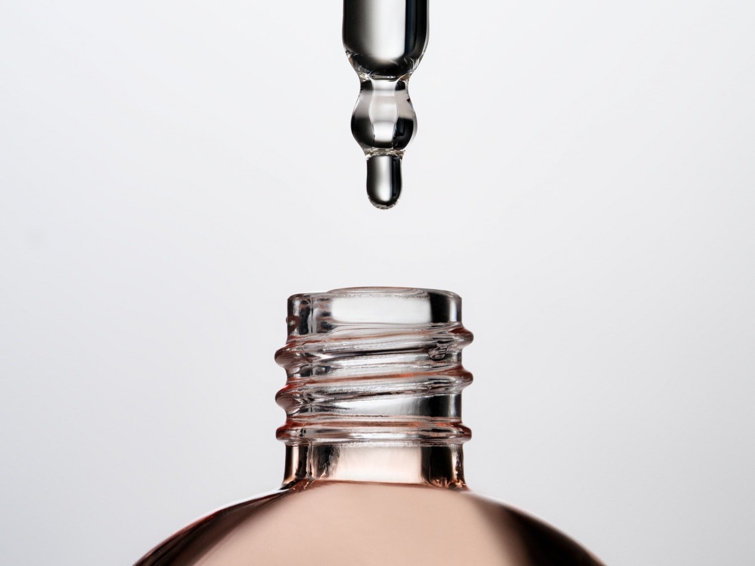 Ein Tropfen eines rosafarbenen Serums fällt aus einer Pipette in den offenen Hals einer Glasflasche, die ebenfalls das rosafarbene Serum enthält.