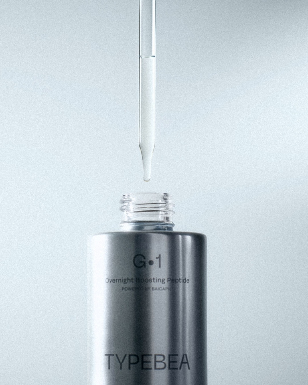 Ein Tropfen des G.1 Overnight Boosting Peptide Serums von TYPEBEA fällt aus einer Pipette in die silberne Flasche, die für eine verbesserte Hautregeneration über Nacht sorgt.