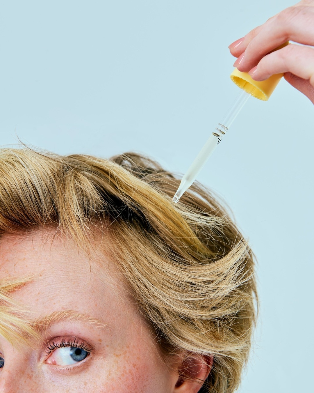 Detail obličeje s blond vlasy, na které se aplikuje sérum pomocí kapátka s žlutým uzávěrem.