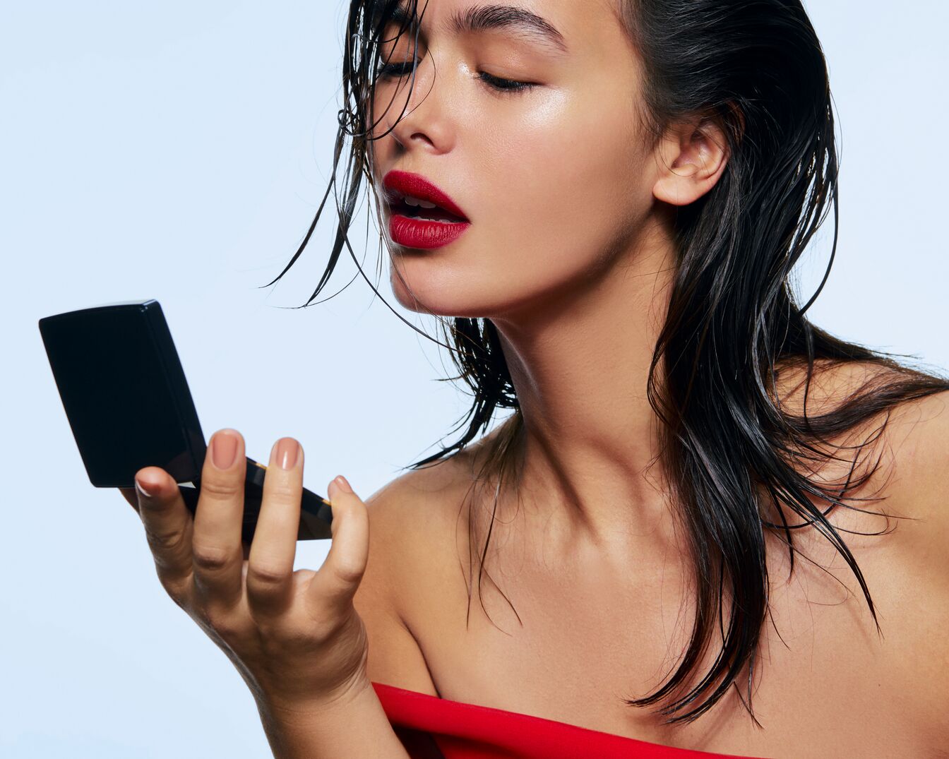 Eine Frau mit nassen, dunklen Haaren und roten Lippen schaut in einen schwarzen Kompaktspiegel, um ihr Make-up zu überprüfen.