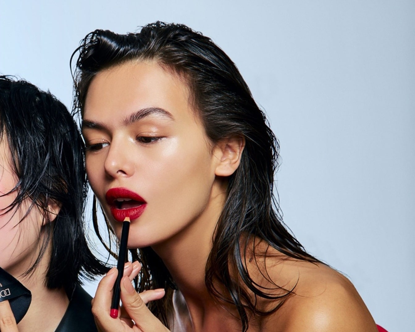 Eine Frau mit glänzender Haut und nassen, dunklen Haaren trägt einen roten Lippenstift mit einem schwarzen Lippenkonturenstift auf.