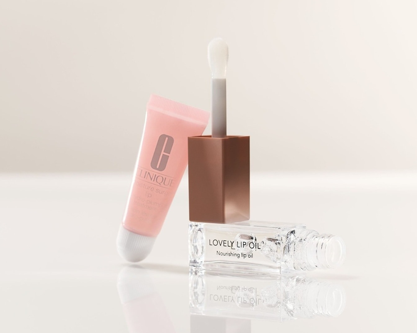 Ein rosa Clinique Moisture Surge Lip Hydro-Plump Treatment Tube, ein Lovely Lip Oil mit braunem Deckel und ein durchsichtiges, nährendes Lippenöl liegen auf einer glänzenden Oberfläche.
