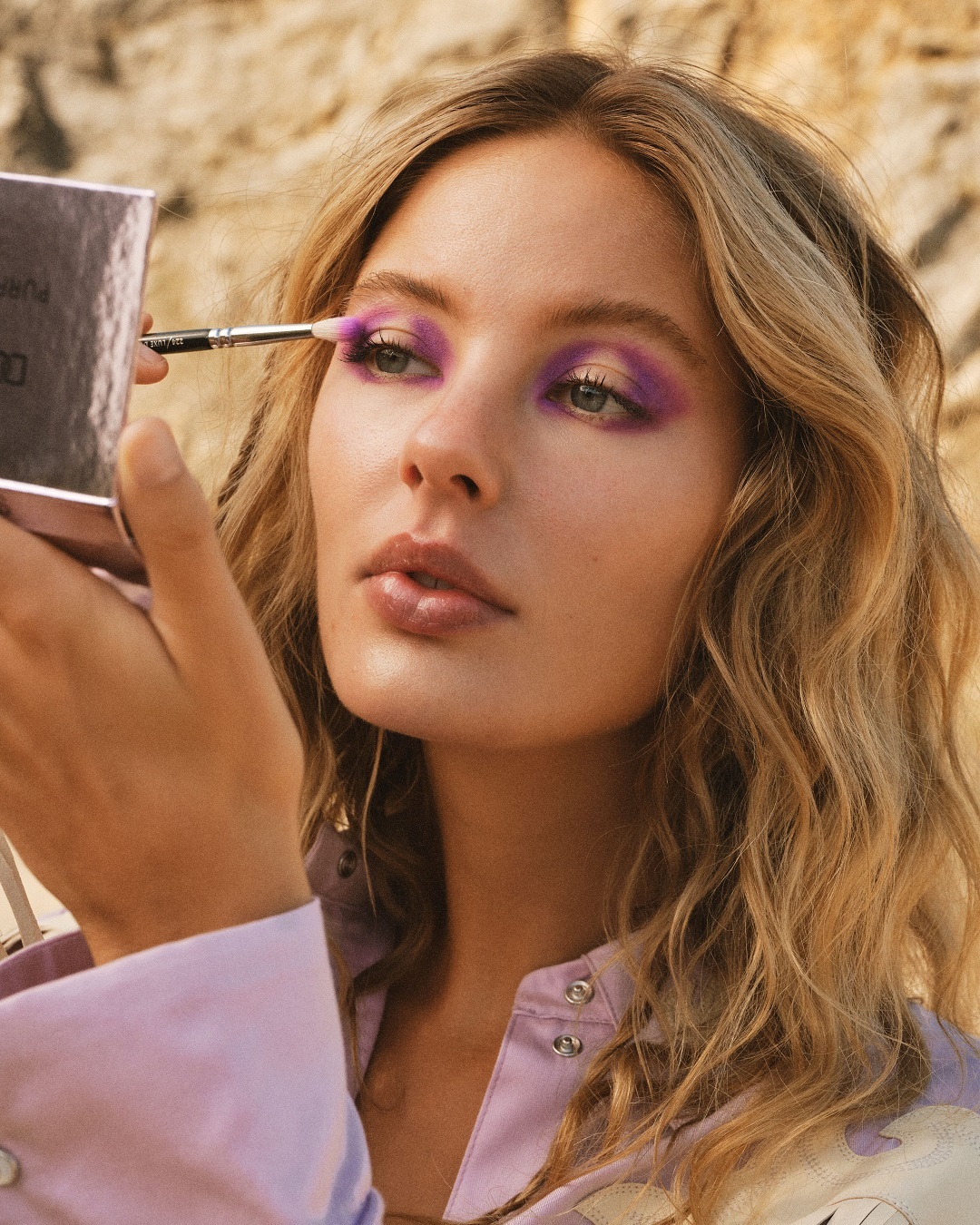 Eine blonde Frau mit gewelltem Haar trägt mit einem Pinsel lila Lidschatten auf und blickt in einen silbernen Spiegel.