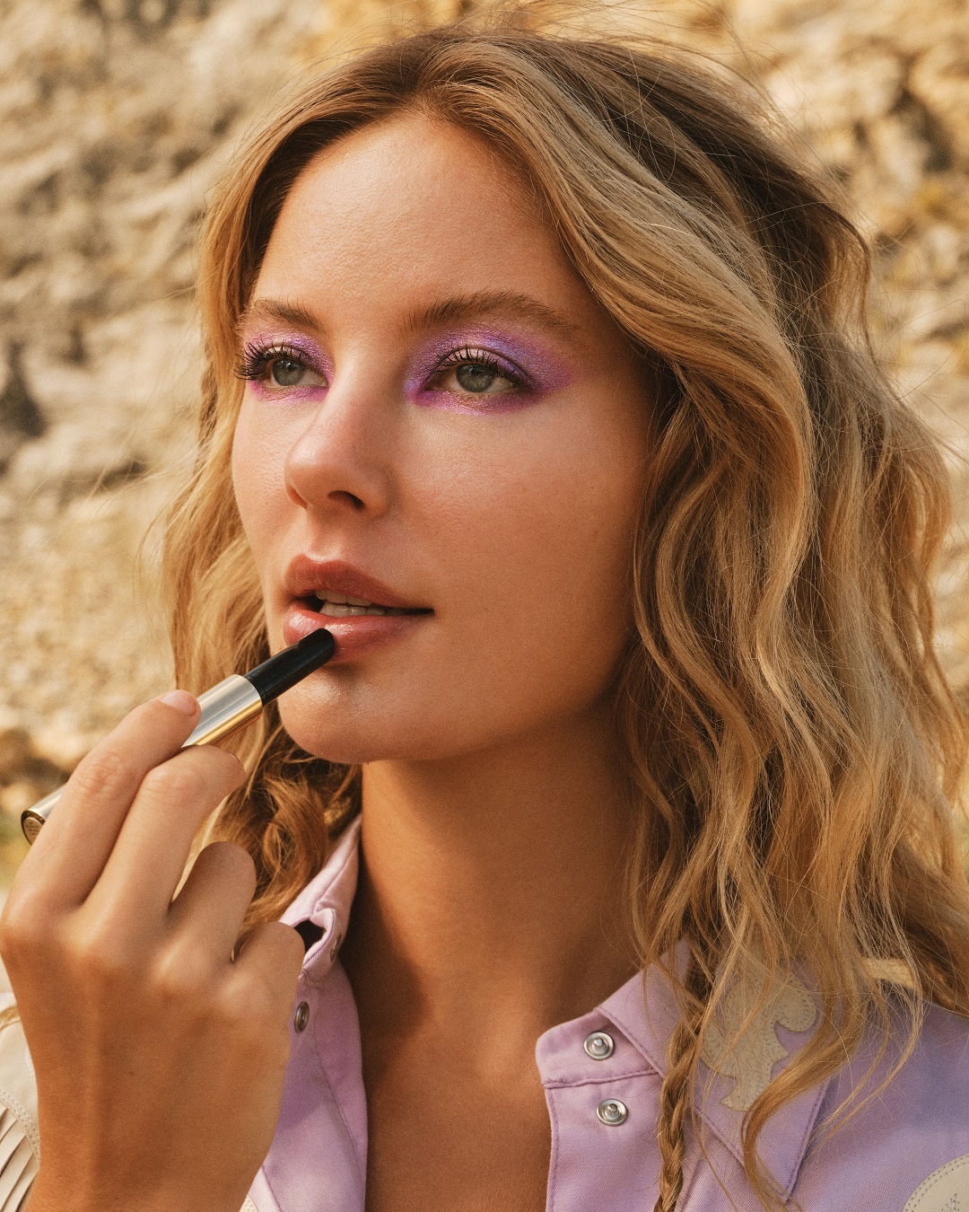 Eine Frau mit gewelltem, blondem Haar trägt einen schwarzen Lippenstift auf, ihr Augen-Make-up ist lila.