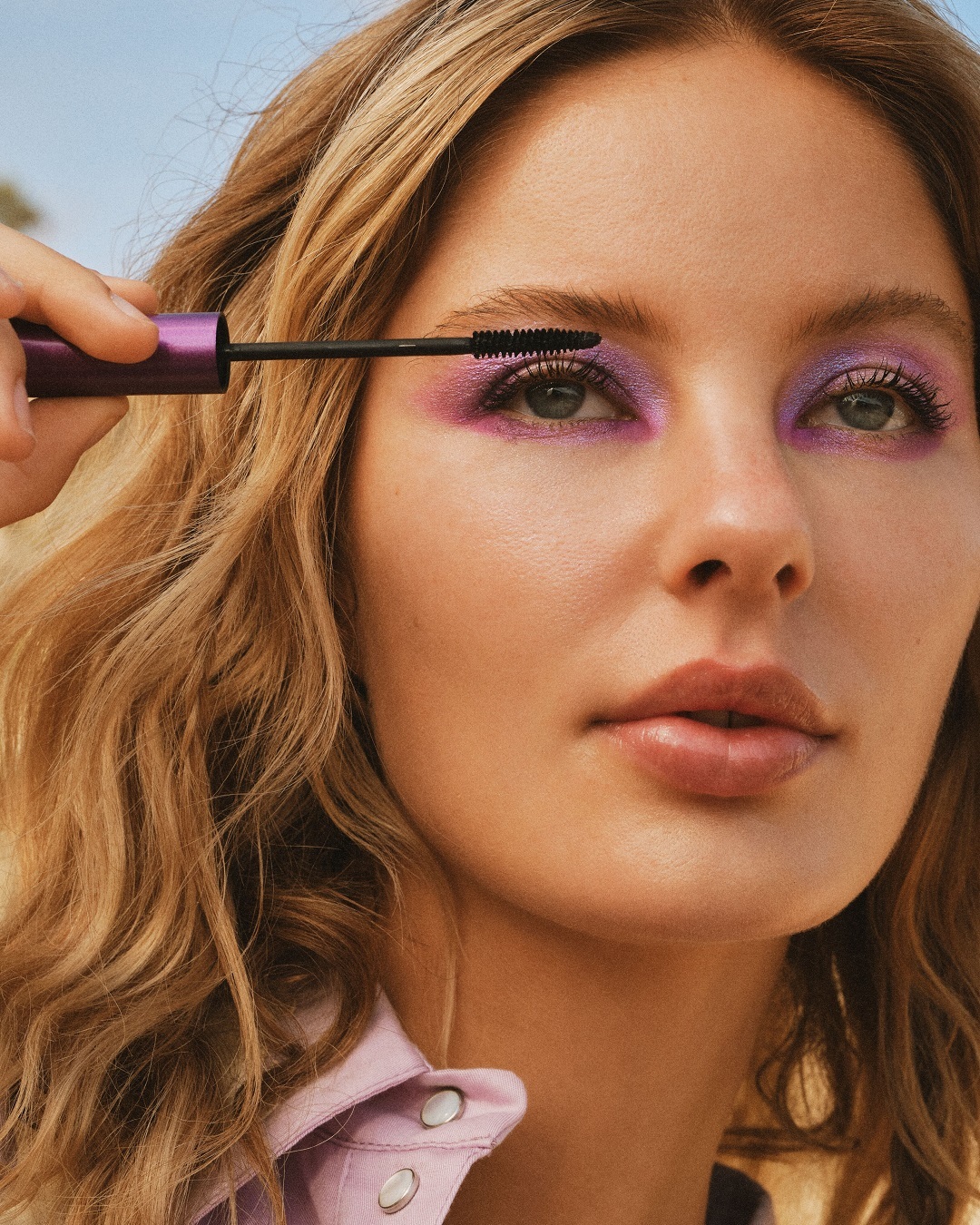 Nahaufnahme einer Frau mit lila Lidschatten, die sich mit einer violetten Mascara die Wimpern tuscht.