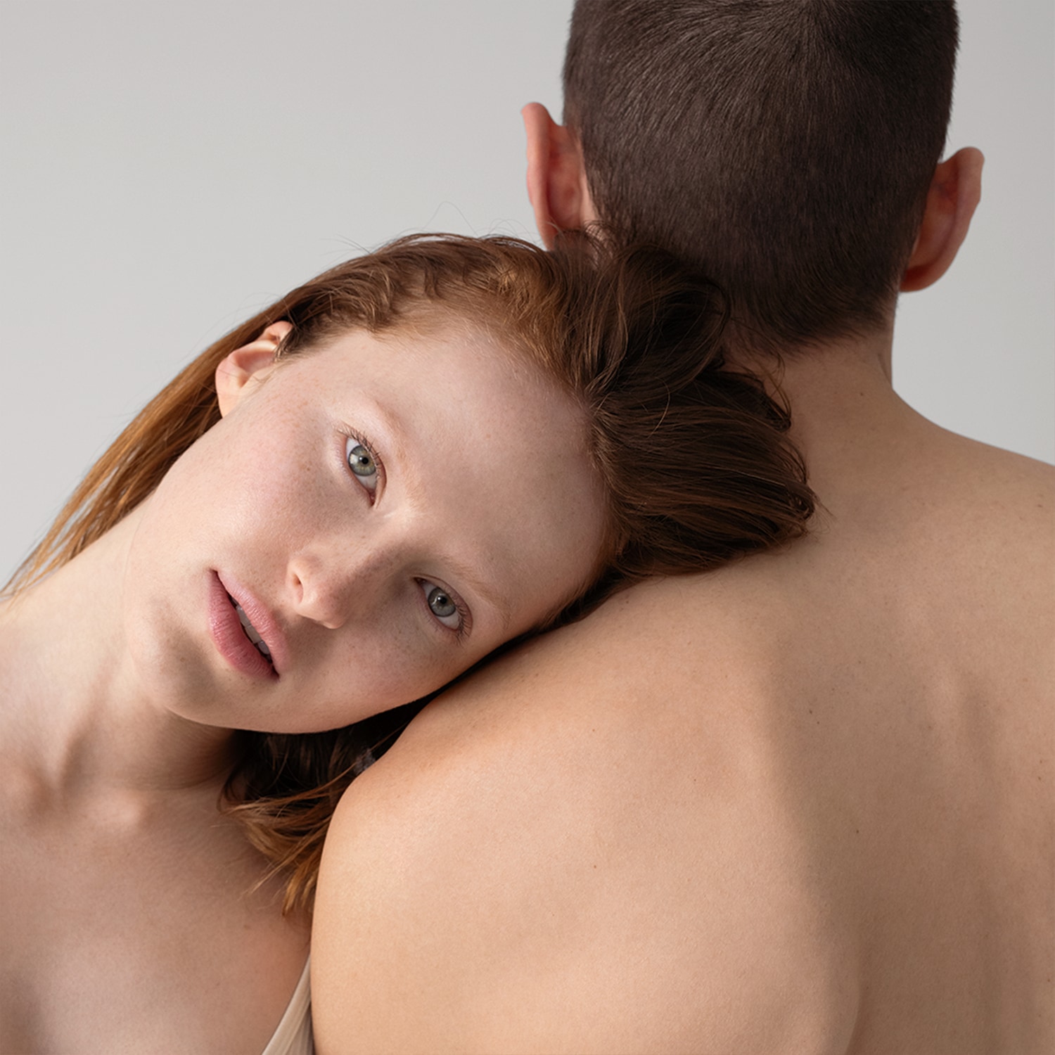 Eine Frau mit roten Haaren und Sommersprossen lehnt ihren Kopf an den Rücken eines Mannes, beide mit heller Haut vor einem neutralen Hintergrund.