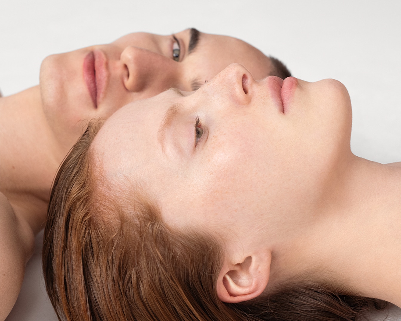 Nahaufnahme von zwei jungen Menschen, die mit geschlossenen Augen auf einem weißen Hintergrund liegen, wobei die Hauttöne und Gesichtszüge im Detail dargestellt sind.