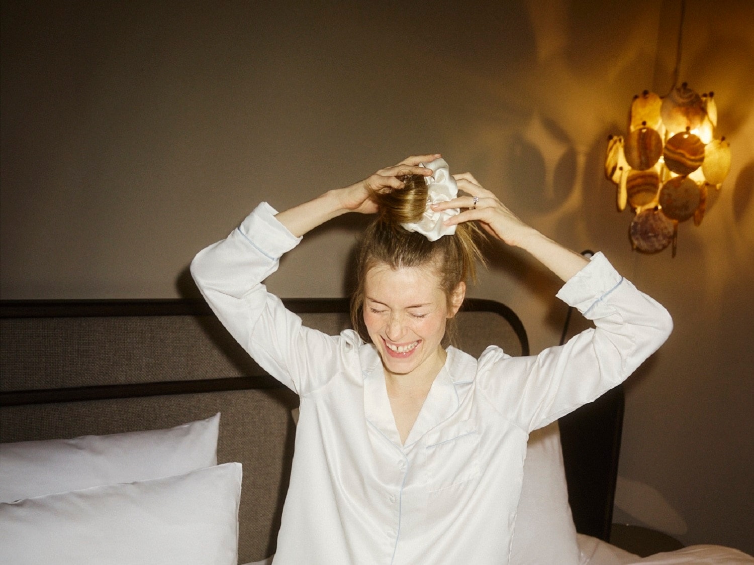 Une femme souriante en pyjama blanc attache ses cheveux avec un chouchou blanc dans une chambre éclairée par une lampe décorative.