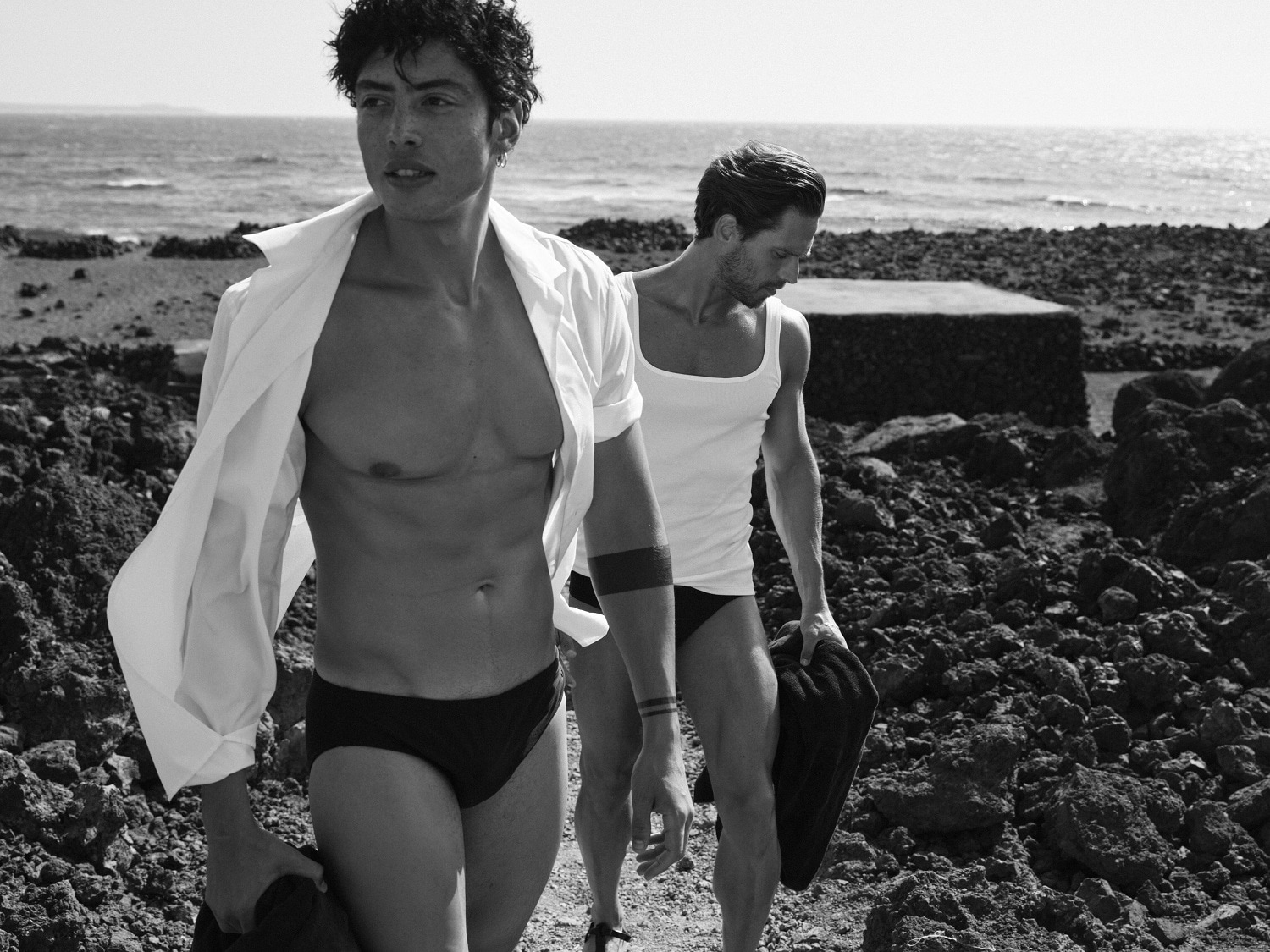 En una playa rocosa en blanco y negro, dos hombres con trajes de baño oscuros y camisetas blancas disfrutan del sol y del mar.