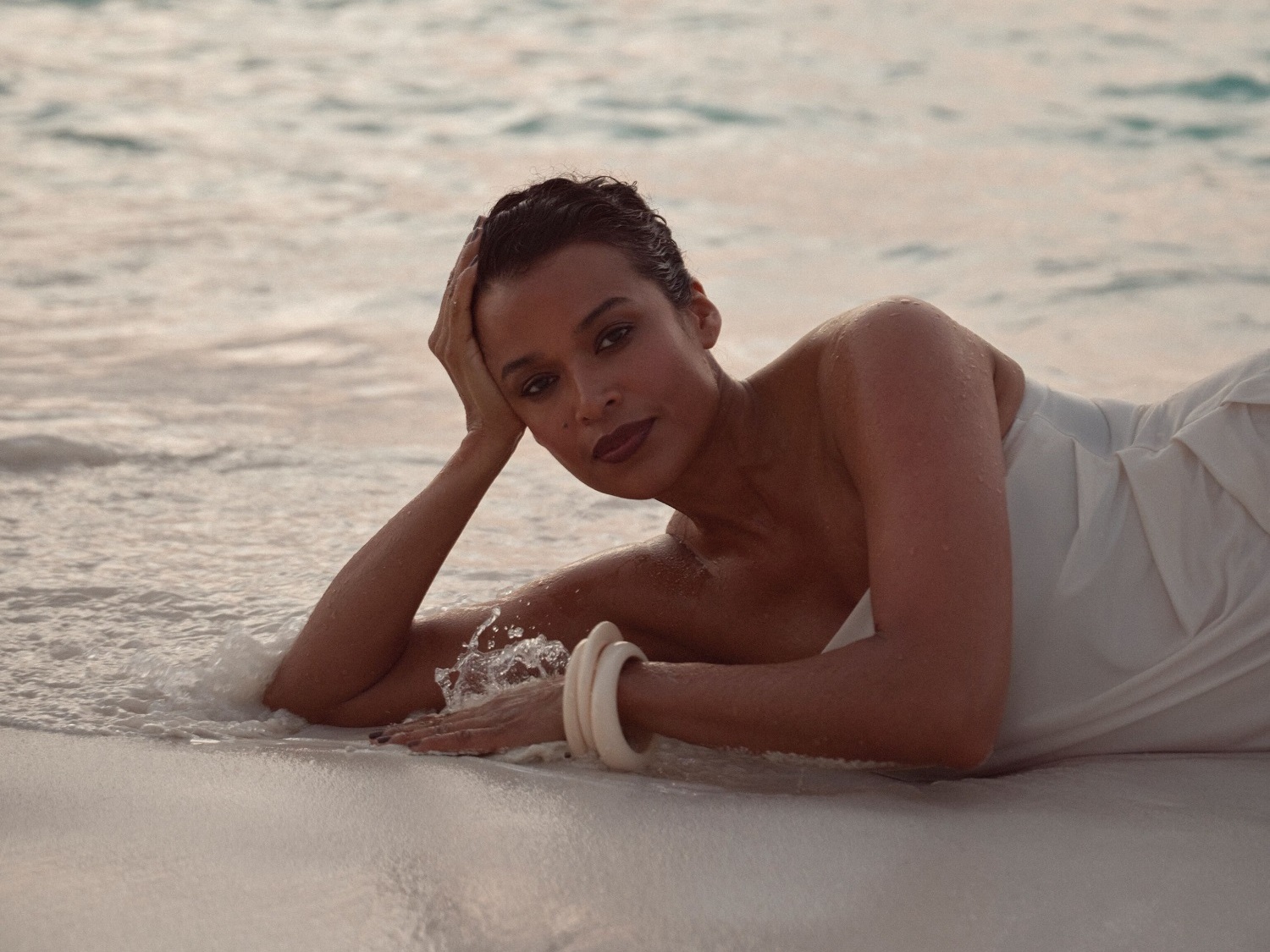 Una mujer con un vestido blanco y un brazalete color crema posa en la playa con una mano en la cabeza y mirando a la cámara.