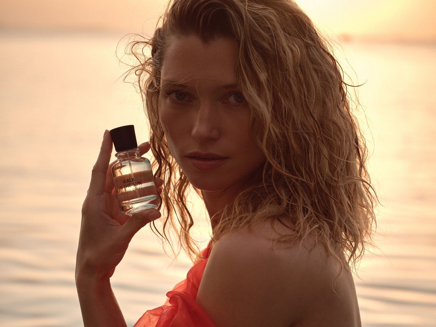Una mujer rubia sostiene un frasco transparente de perfume "Walk in the Sun" con tapón negro, resaltando su aroma veraniego y fresco.