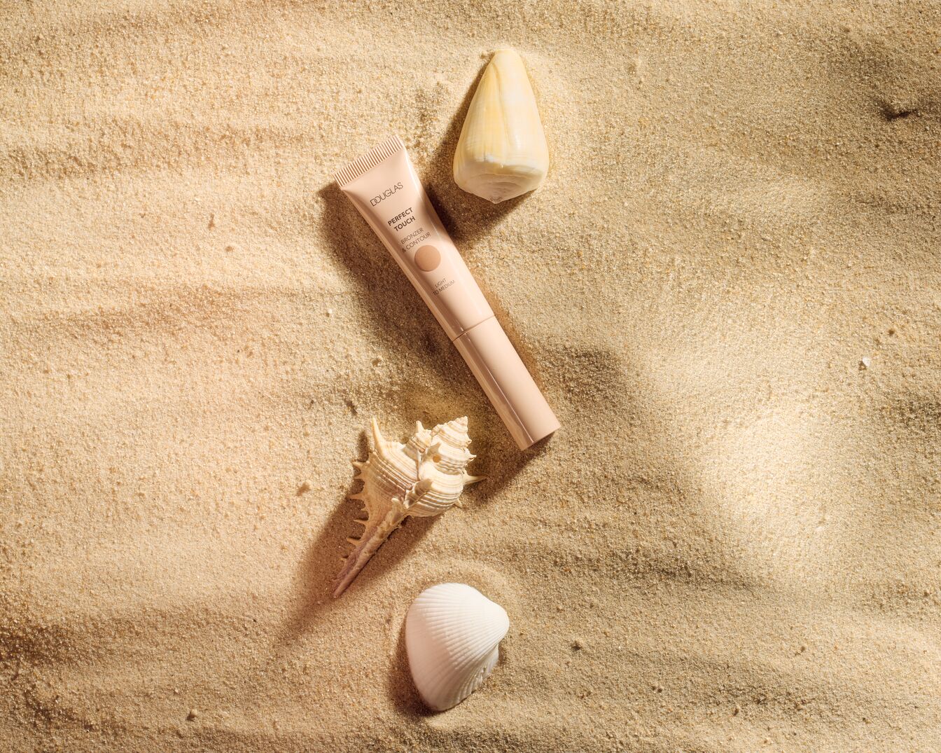 Auf Sand liegt eine Tube Douglas Perfect Touch Bronzer & Contour in hellem bis mittlerem Farbton, umgeben von Muscheln.