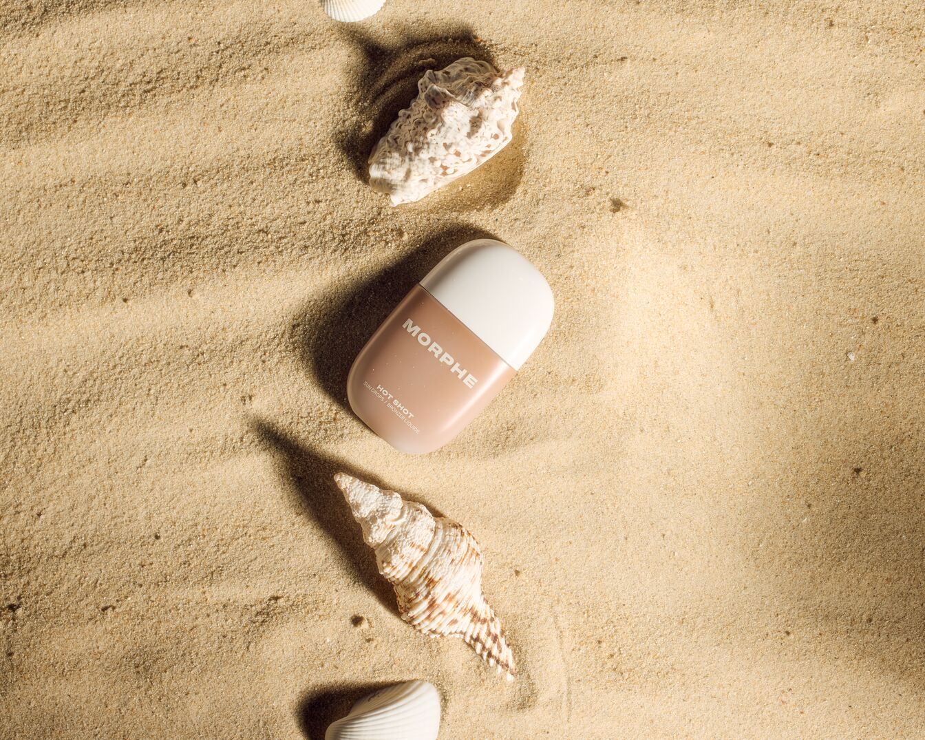 Morphe Hot Shot Sonnenglanz-Bronzer auf Sand mit Muscheln, die eine sommerliche Atmosphäre für einen strahlenden Teint erzeugen.