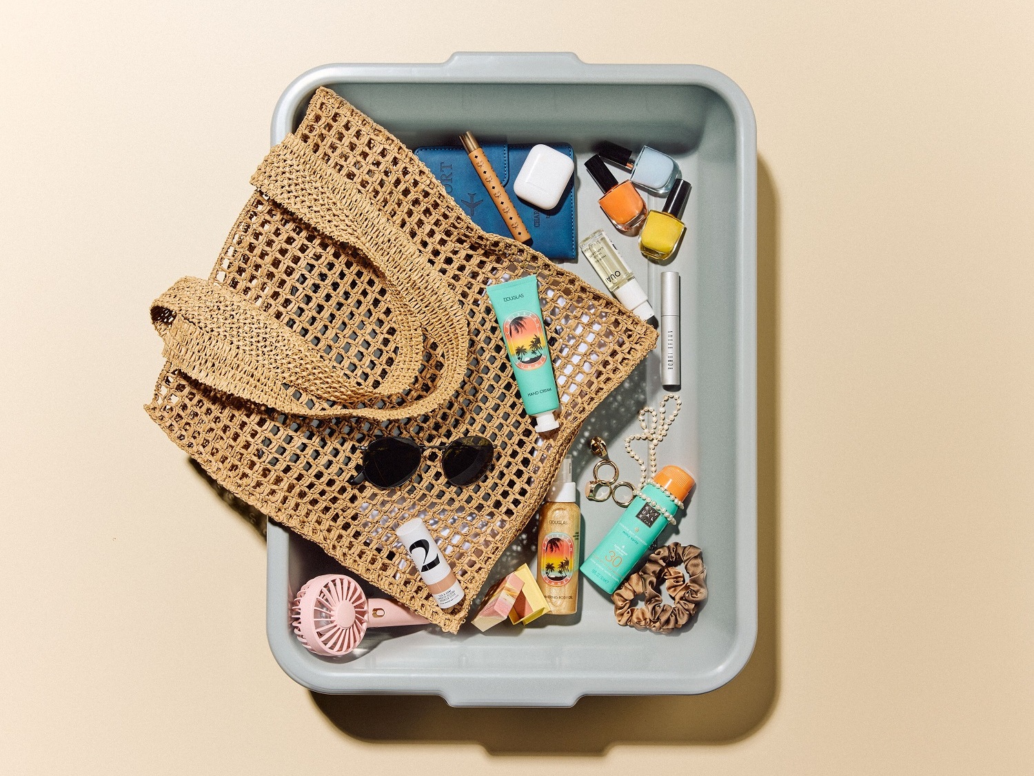 Draufsicht auf eine graue Schale mit einer gehäkelten Strandtasche, Sonnenbrille, einem rosa Ventilator, Kosmetika und Accessoires, die für einen Tag am Strand bereit sind.