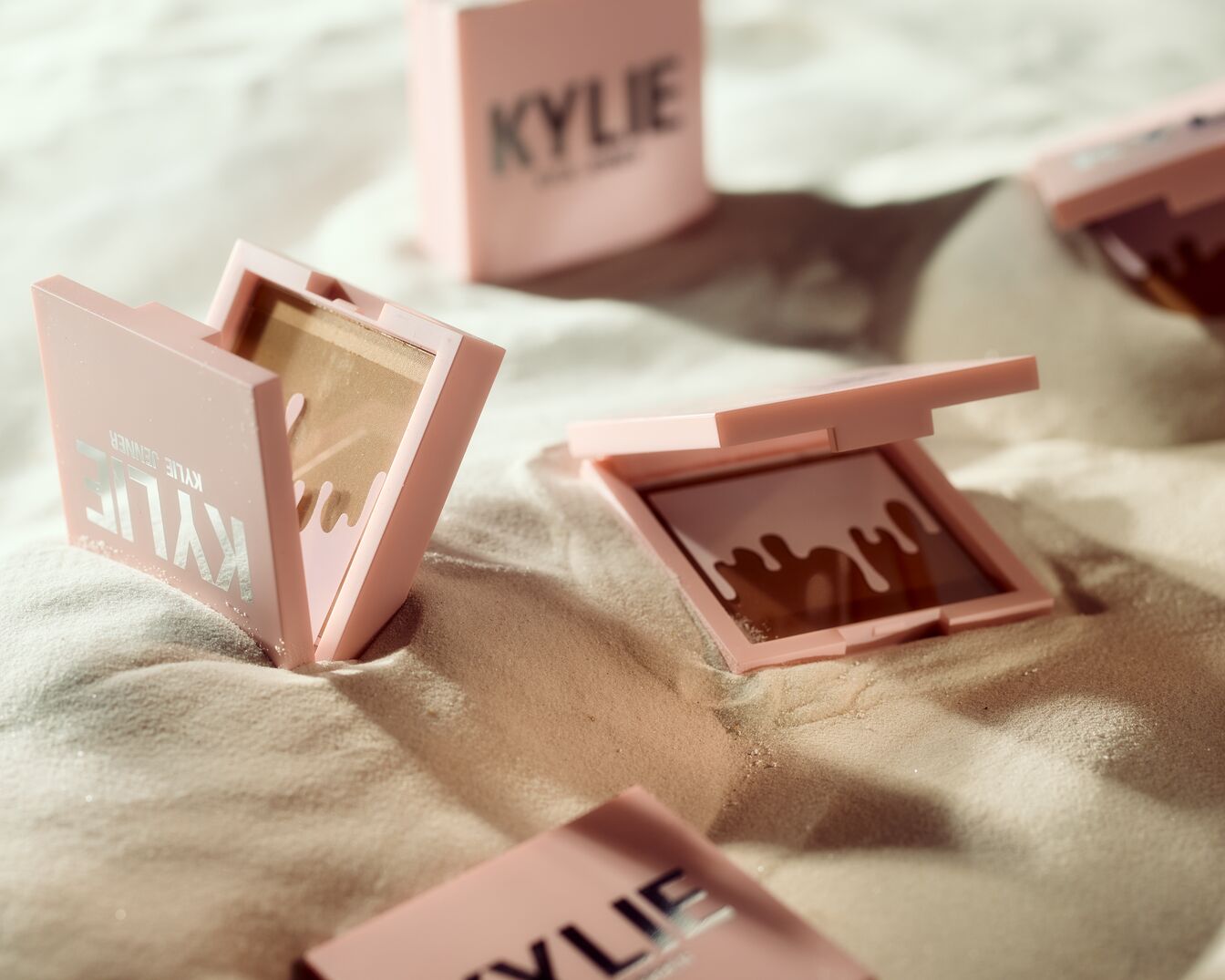 Nahaufnahme von Kylie Jenner Kompaktpuder in hellrosa Verpackung, eingebettet in Sand, wobei die Textur und Farbe des Produkts hervorgehoben werden.
