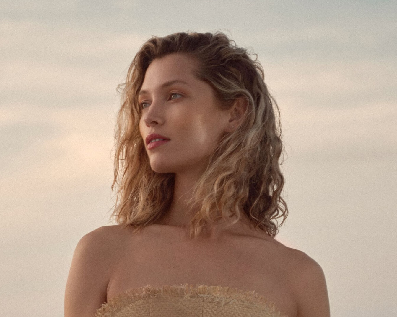 Eine Frau mit gewelltem, blondem Haar und einem schulterfreien Oberteil aus Bast blickt vor einem hellen Himmel verträumt in die Ferne.