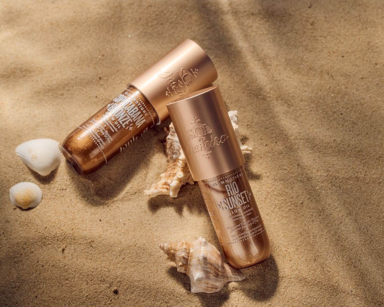 Two Sol de Janeiro glow oil bottles, one labeled 'Copacabana Bronze' and the other 'Rio Sunset,' rest on a sandy surface with scattered seashells, promising a radiant, sun-kissed shimmer for body and face.