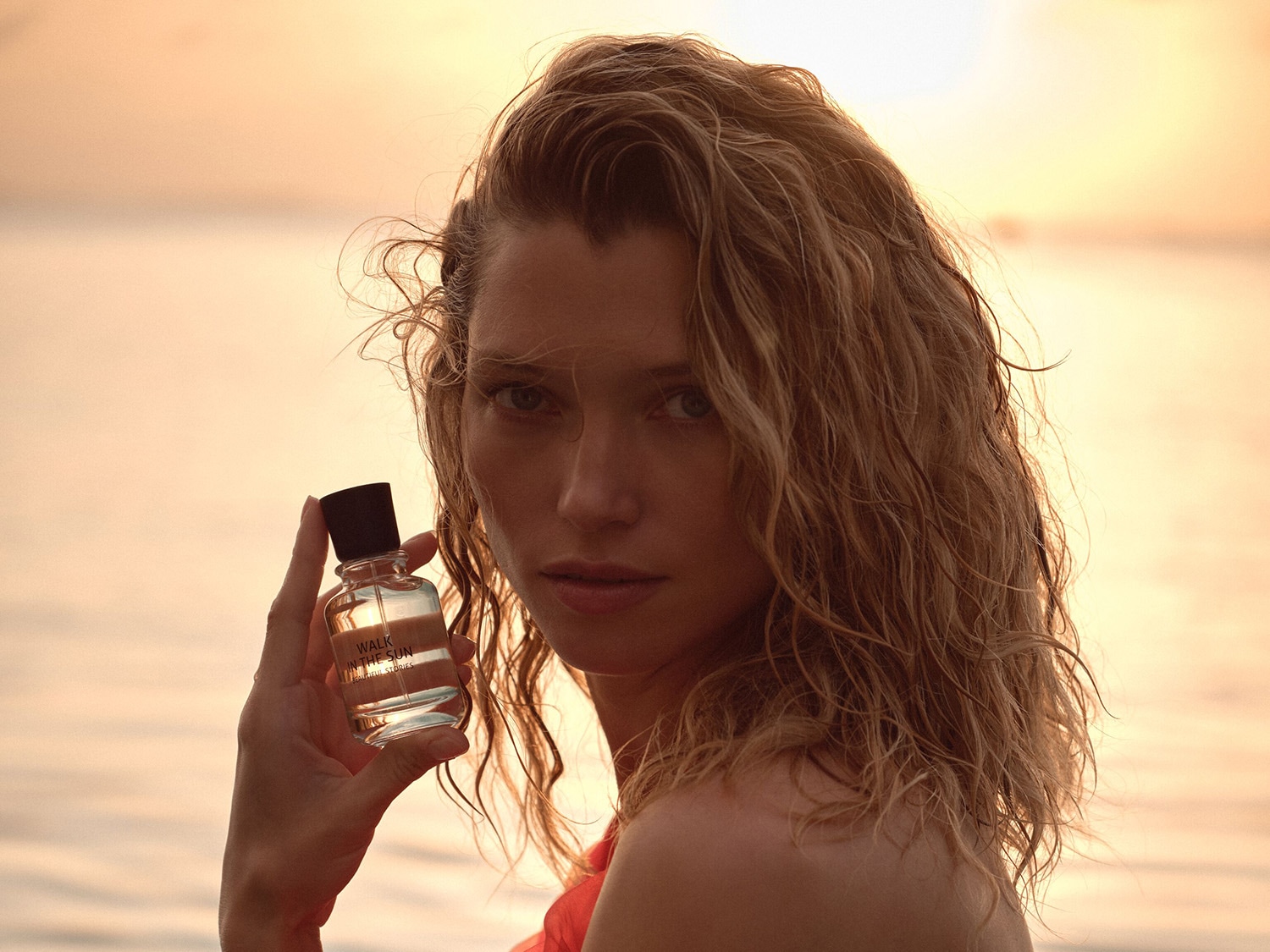 Eine blonde Frau mit nassen Haaren hält eine durchsichtige Parfümflasche mit der Aufschrift „Walk in the Sun“ vor einem orangefarbenen Hintergrund.