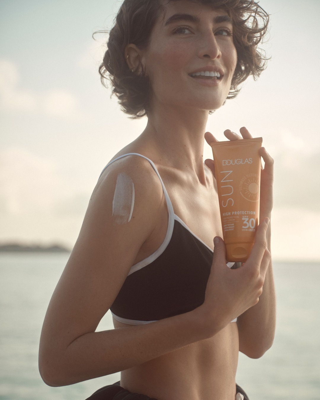 Une femme souriante tient un tube orange de crème solaire Douglas Sun Haute Protection FPS 30, avec une trace de crème sur son épaule.