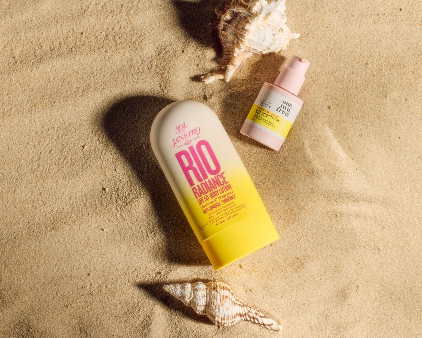 Voici un écran solaire minéral SPF 50 Rio Radiance de Sol de Janeiro, à côté d'un écran solaire quotidien One Two Free, le tout sur du sable avec des coquillages.