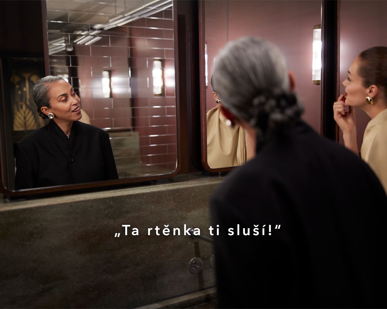 Two women in a bathroom, one applying red lipstick while the other admires her reflection, with the phrase "Ta rtěnka ti sluší!" suggesting the lipstick looks great.