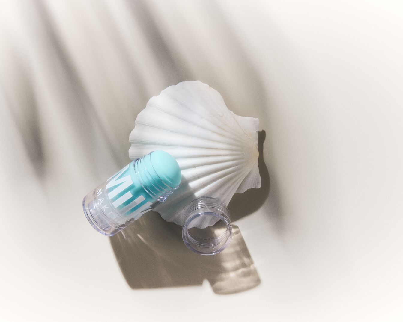 Barra de maquillaje Milk con tapa azul claro, apoyada sobre una concha blanca, con su tapa transparente al lado, sobre una superficie blanca iluminada por la luz del sol.