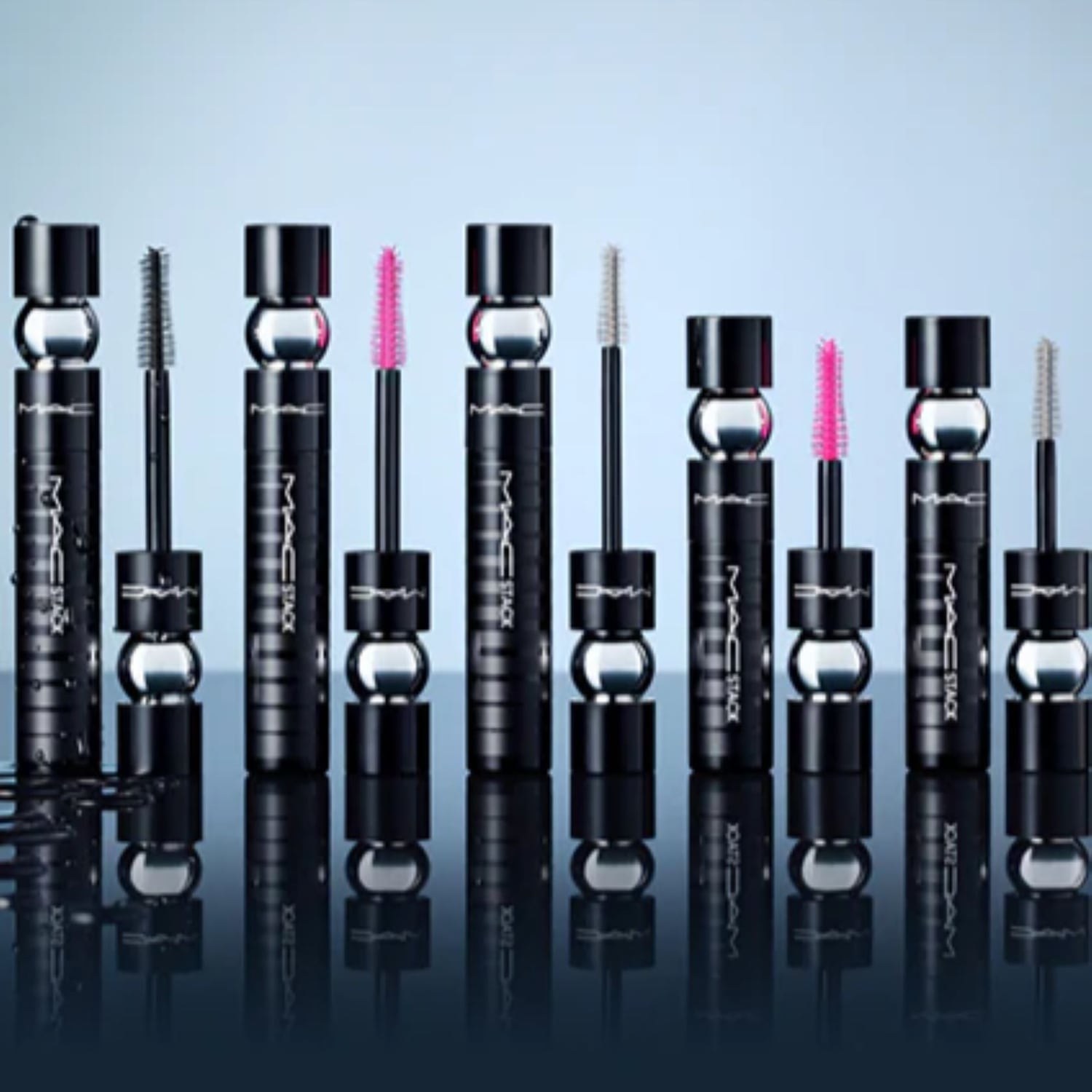 Eine Reihe von MAC Stack Mascaras in schwarzen Tuben mit silbernen Akzenten und verschiedenen Bürsten, die auf einer glänzenden Oberfläche stehen.