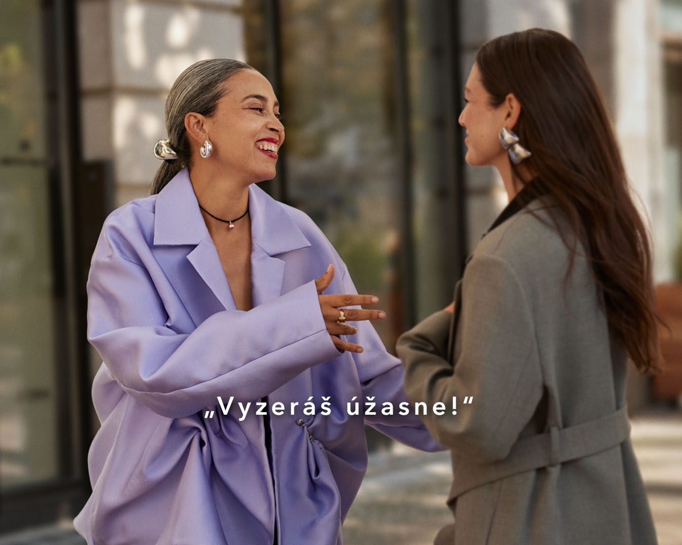 Dve ženy sa usmievajú a rozprávajú, jedna má na sebe fialový kabát a druhá sivý kabát, vyzerajú úžasne!