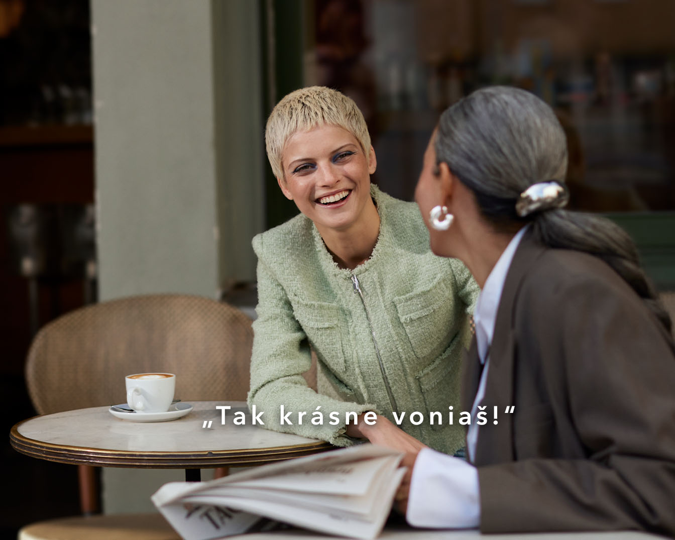 Two women are sitting at a table outside a cafe, one with short blonde hair in a green jacket, smiling at the other with gray hair in a brown suit, with the text "Tak krásne voniaš!"