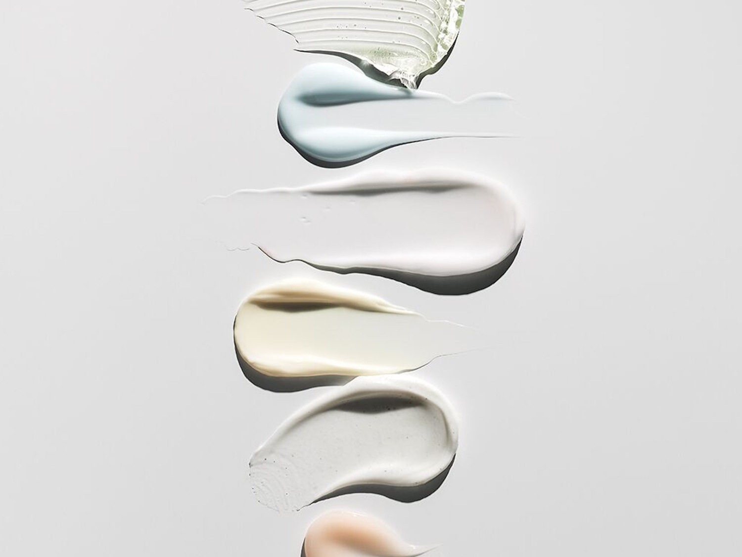 Na białym tle widoczne są próbki różnych kosmetyków o kremowej konsystencji i zróżnicowanej kolorystyce, od przezroczystej, przez błękitną, białą, żółtą, szarą, aż po brzoskwiniową.