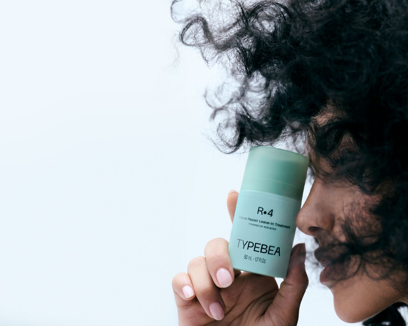 Kobieta z kręconymi włosami trzyma w dłoni zieloną butelkę TYPEBEA R.4 Intense Repair Leave-In Treatment o pojemności 50 ml, z widocznym napisem „POWERED BY RDS BOND”.