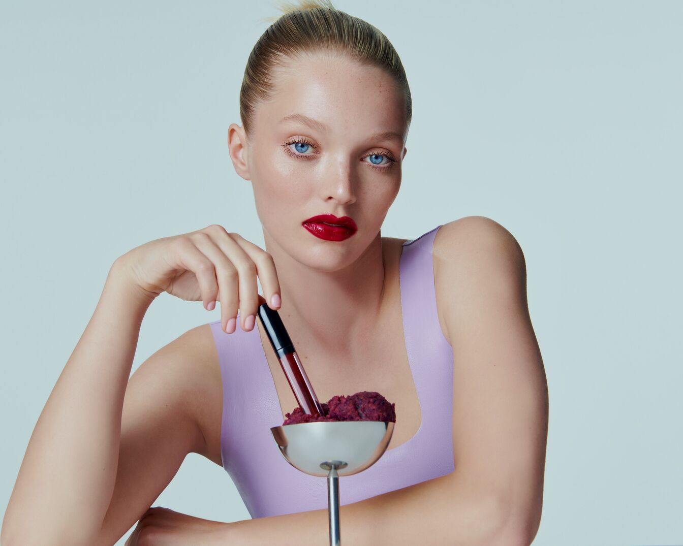 Een vrouw met blauwe ogen en rode lippenstift houdt een zilveren beker vast met een rode vloeistof en een zwarte dop.