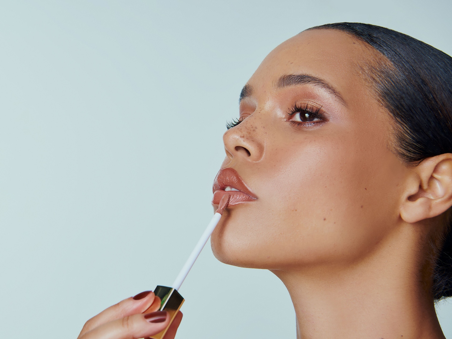 Een vrouw brengt glanzende nude lipgloss aan met een applicator met gouden dop, voor een subtiele en stralende look.