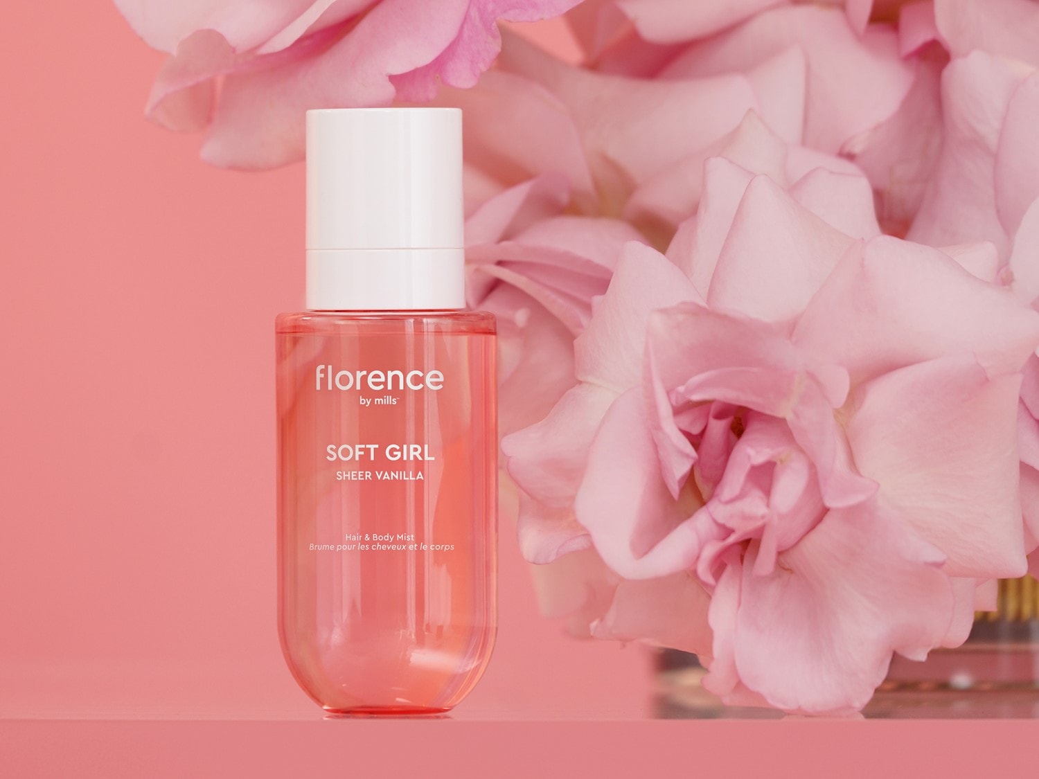Rožiniame fone matomas „Florence by Mills Soft Girl Sheer Vanilla“ plaukų ir kūno dulksnos buteliukas su baltu dangteliu, apsuptas švelniai rožinių gėlių.