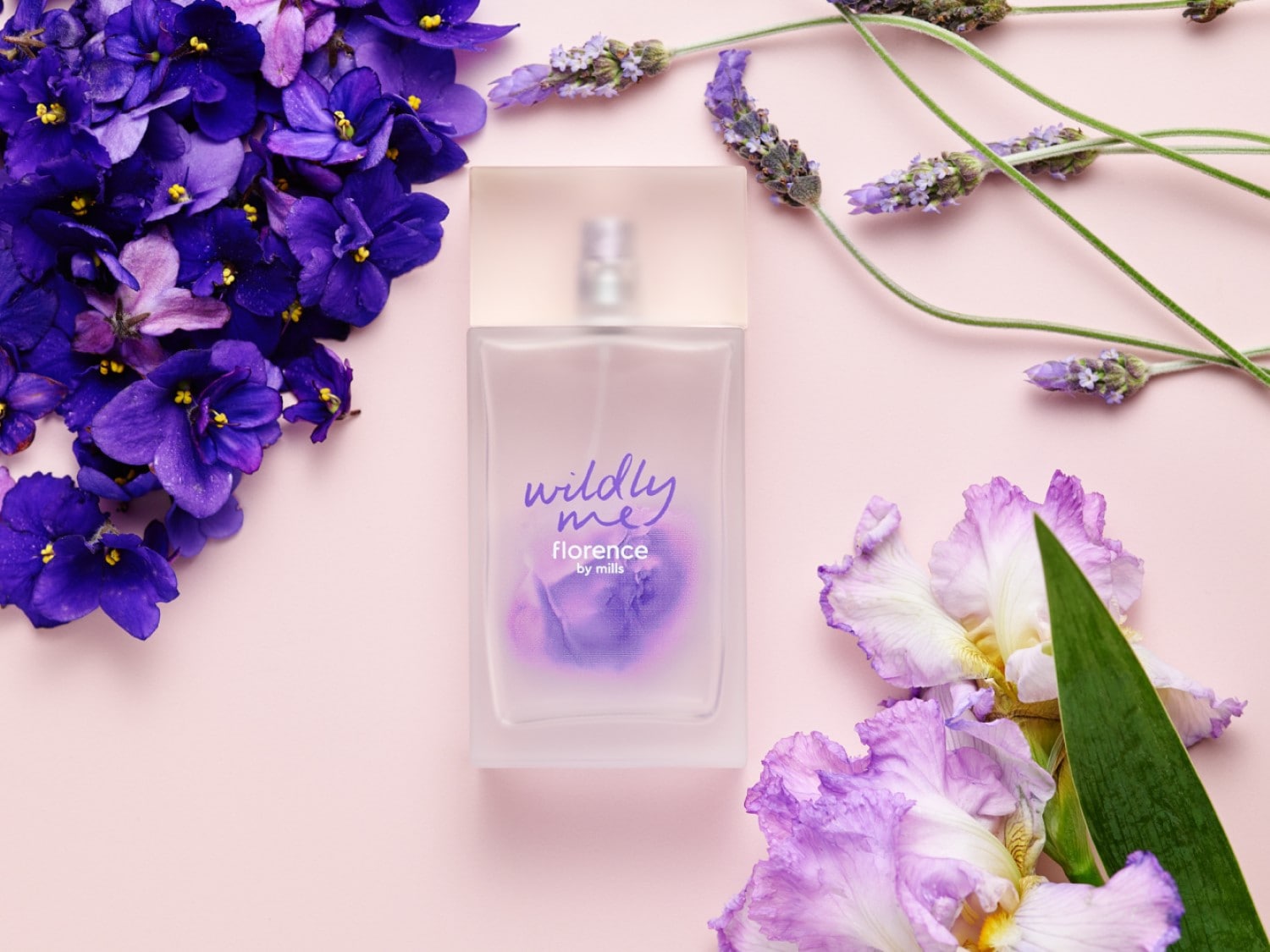 Švelniai rožiniame fone pavaizduotas „Florence by Mills“ „Wildly Me“ aromatas su violetinėmis gėlėmis ir levandomis.