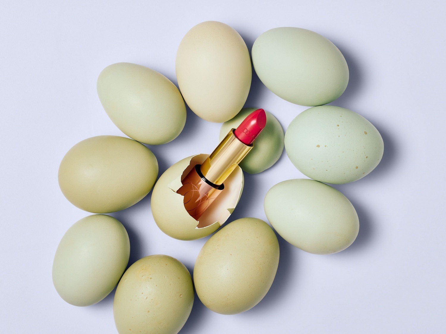 Rodeado de huevos color aguacate, un labial rojo emerge de un cascarón roto, simbolizando un nuevo comienzo en el mundo de la belleza.