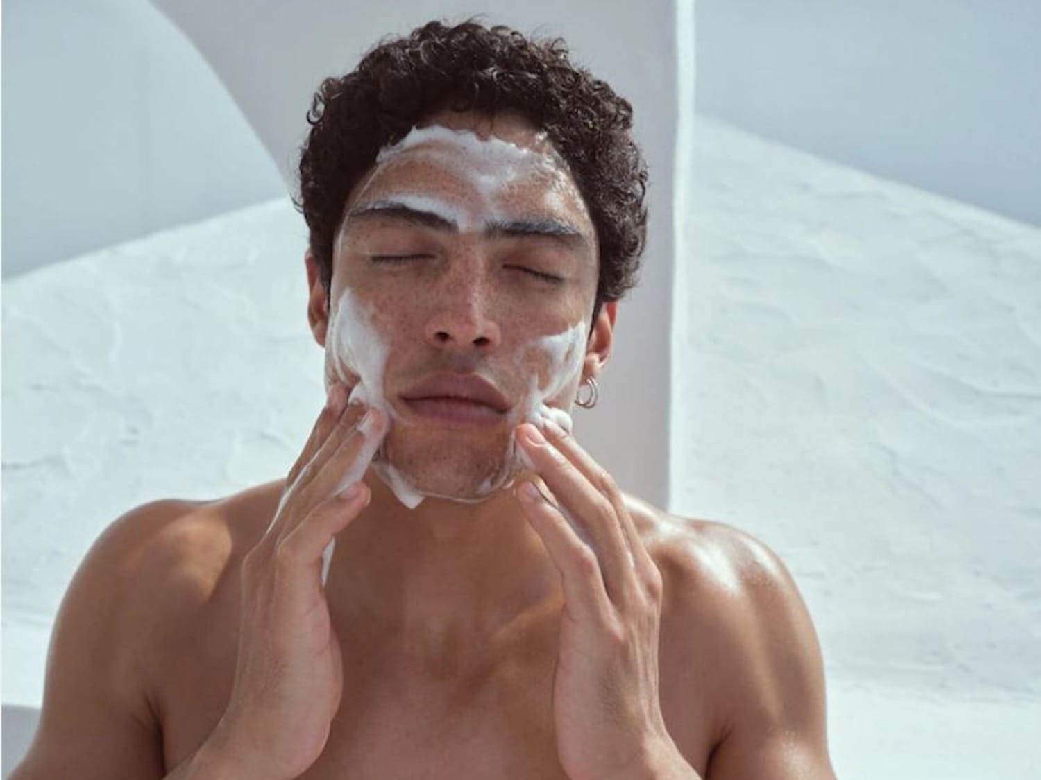 Un homme aux cheveux bouclés, les yeux fermés, se nettoie le visage avec une mousse blanche, créant une sensation de fraîcheur et de pureté.