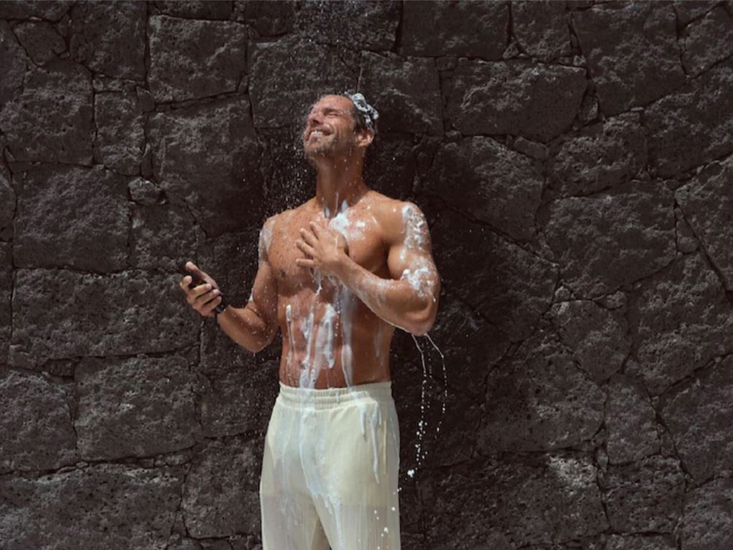 Un homme souriant, torse nu et en pantalon blanc, se douche avec un téléphone à la main, devant un mur de pierre sombre.