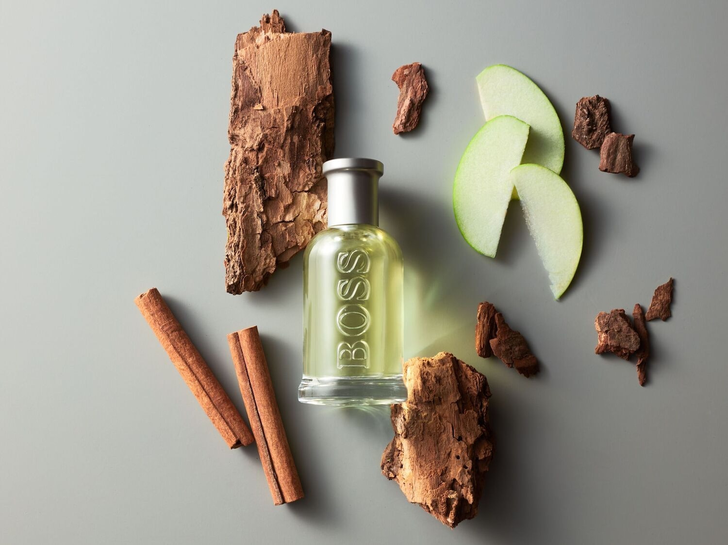 Viršutinis vaizdas ant pilko paviršiaus, kuriame yra „Boss“ kvepalų buteliukas, du cinamono pagaliukai, trys obuolių griežinėliai ir medžio žievės gabalėliai.