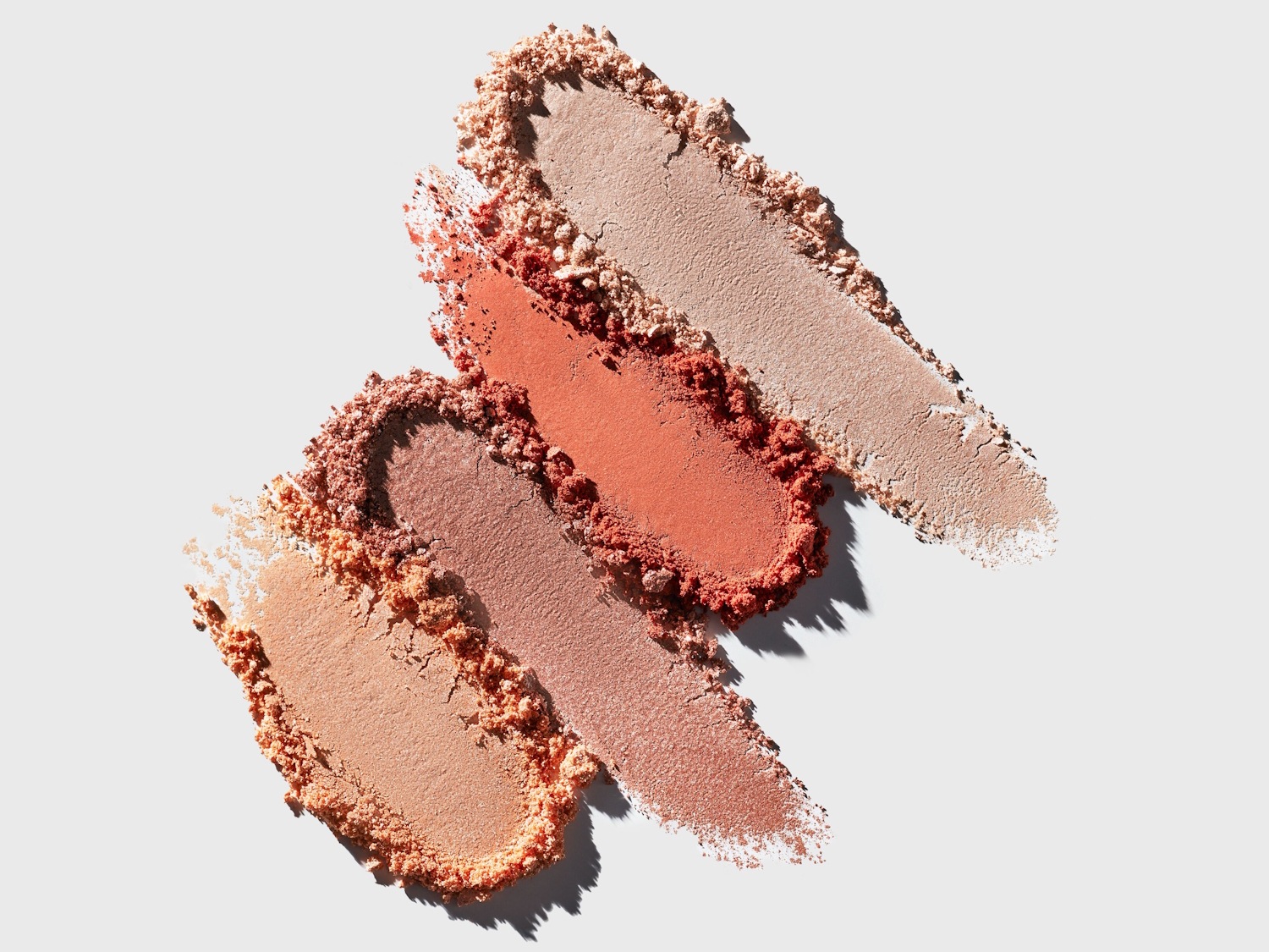 Vier Swatches von Puder-Make-up in verschiedenen Hauttönen, von hellem Beige über Rosé bis hin zu einem warmen Terrakotta, sind diagonal auf einem weißen Hintergrund angeordnet.