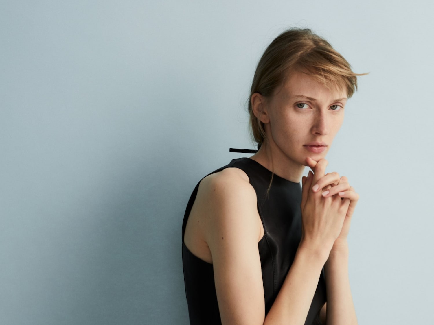 Een vrouw met blond haar en een zwarte top poseert tegen een lichtblauwe achtergrond, haar handen gevouwen onder haar kin in een nadenkende pose.