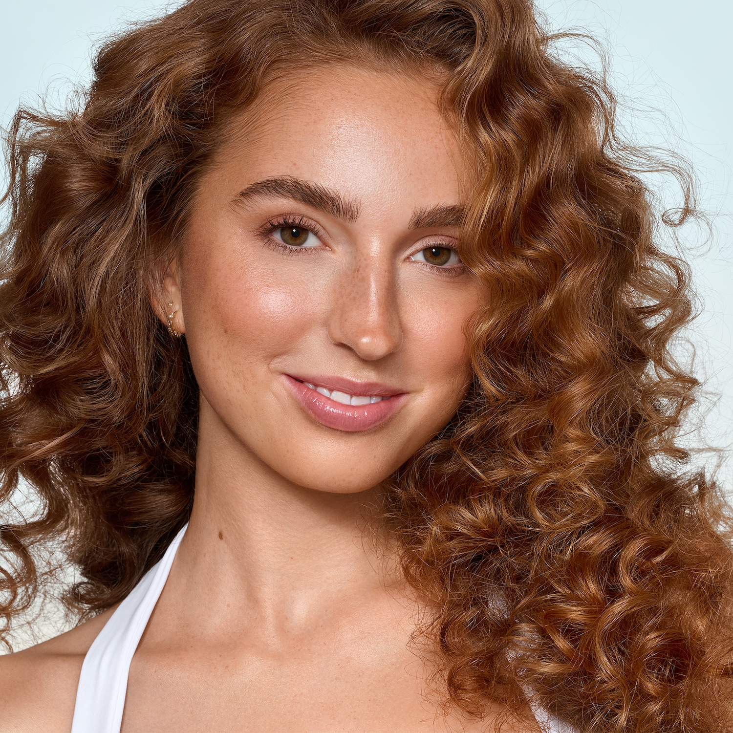 Portrait d'une jeune femme souriante aux cheveux roux bouclés, au teint clair et aux yeux marron, portant un haut blanc.